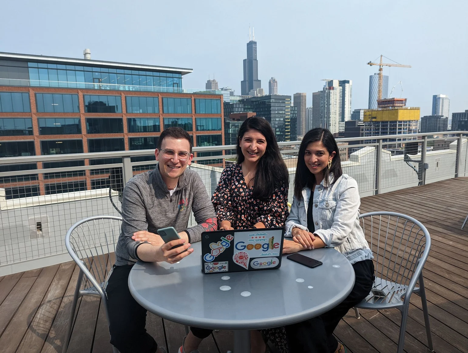 Three people smiling at a rooftop table with laptops and phones at Google Chicago office